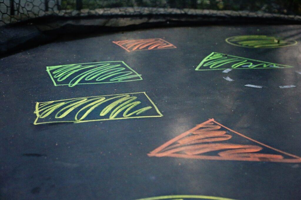 Child jumping on a backyard trampoline decorated with brightly colored sidewalk chalk shapes like red circles and blue triangles, spaced apart for the Trampoline Sidewalk Chalk Learning Game for Kids. A perfect outdoor summer trampoline activity.