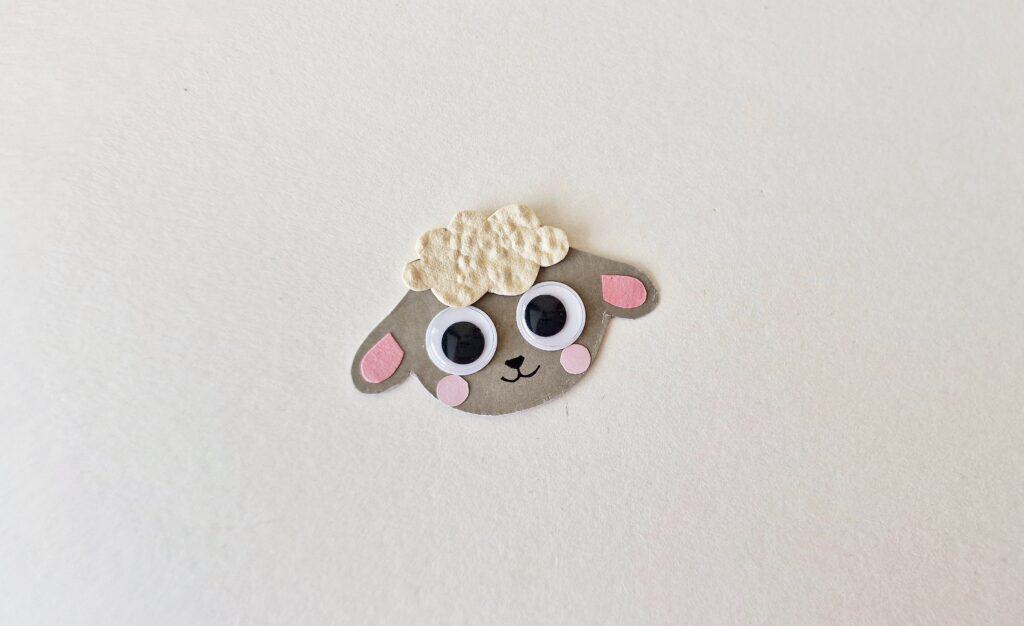 Close-up image showing a cutout of a sheep's head made from colored craft paper. A pair of hands is gluing small pink inner ear pieces onto the larger ear shapes of the sheep’s head. The craft is placed on a clean, flat surface with other paper pieces visible nearby. This step demonstrates adding detail to the ears in the handprint sheep craft for toddlers and preschoolers.








