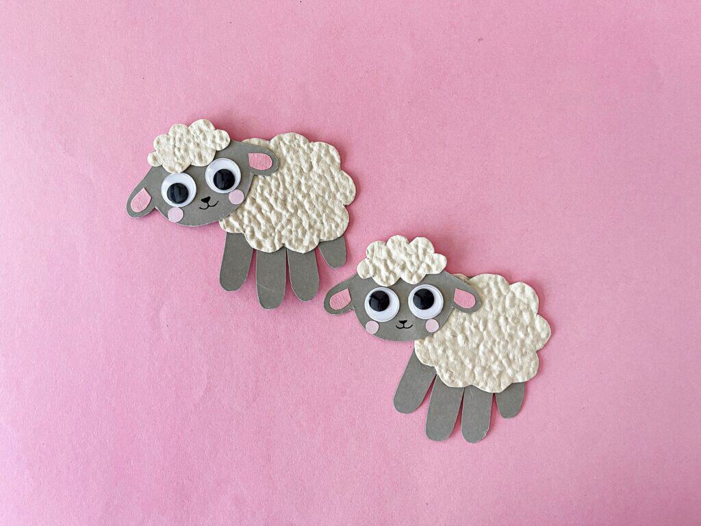 Finished handprint sheep craft for toddlers and preschoolers made with colored craft paper, embossed white paper, googly eyes, and a black sharpie. The sheep's head is glued to the thumb of the handprint, decorated with pink blush and fluffy wool texture. Ideal for farm animal preschool activities, baby animals crafts, and classroom art displays.

