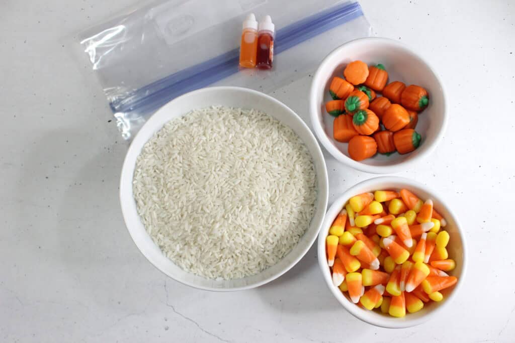 Candy corn sensory bin supplies for toddlers and preschoolers including uncooked rice in Ziploc bags, orange and yellow food dye, stackable sensory bin container, candy corn with pumpkins, measuring tools, baking tray, Halloween figurines, and sensory play tools for fun fall sensory activities and candy corn science.

