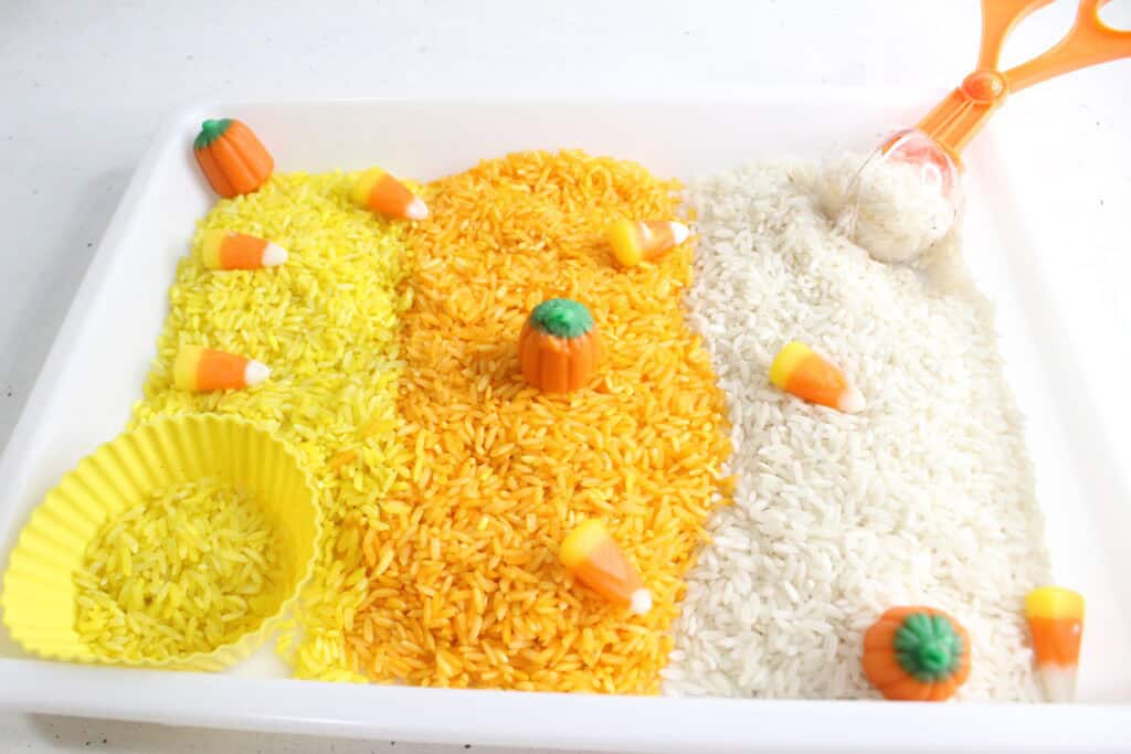 Candy corn sensory bin filled with layers of orange and yellow dyed rice, candy corn with pumpkins, and Halloween-themed figurines for toddlers. Perfect for fall sensory activities, preschool science experiences, and hands-on Halloween candy experiments. Encourages fine motor play and imaginative learning.

