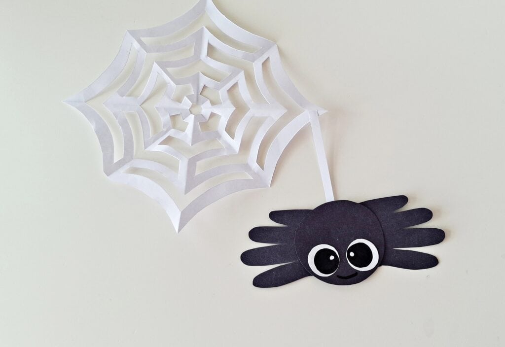 Handprint Spider Steps (14)
Cut a thin strip of white paper to act like “web silk.” Glue one end of the strip to the top of the spider’s head, and the other end to the middle of your folded spider web craft. 